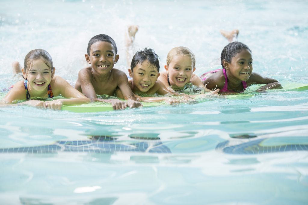 A multi-ethnic group of elementary age children are swimming on their kick boards during a swimming lesson. They are smiling and looking at the camera.